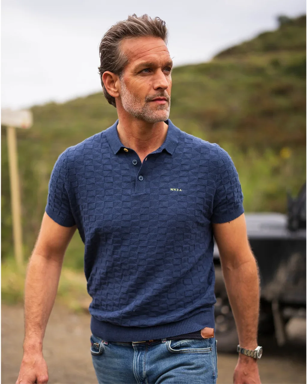 Man in a blue textured polo shirt and jeans walks outdoors, with greenery and a cloudy sky in the background.