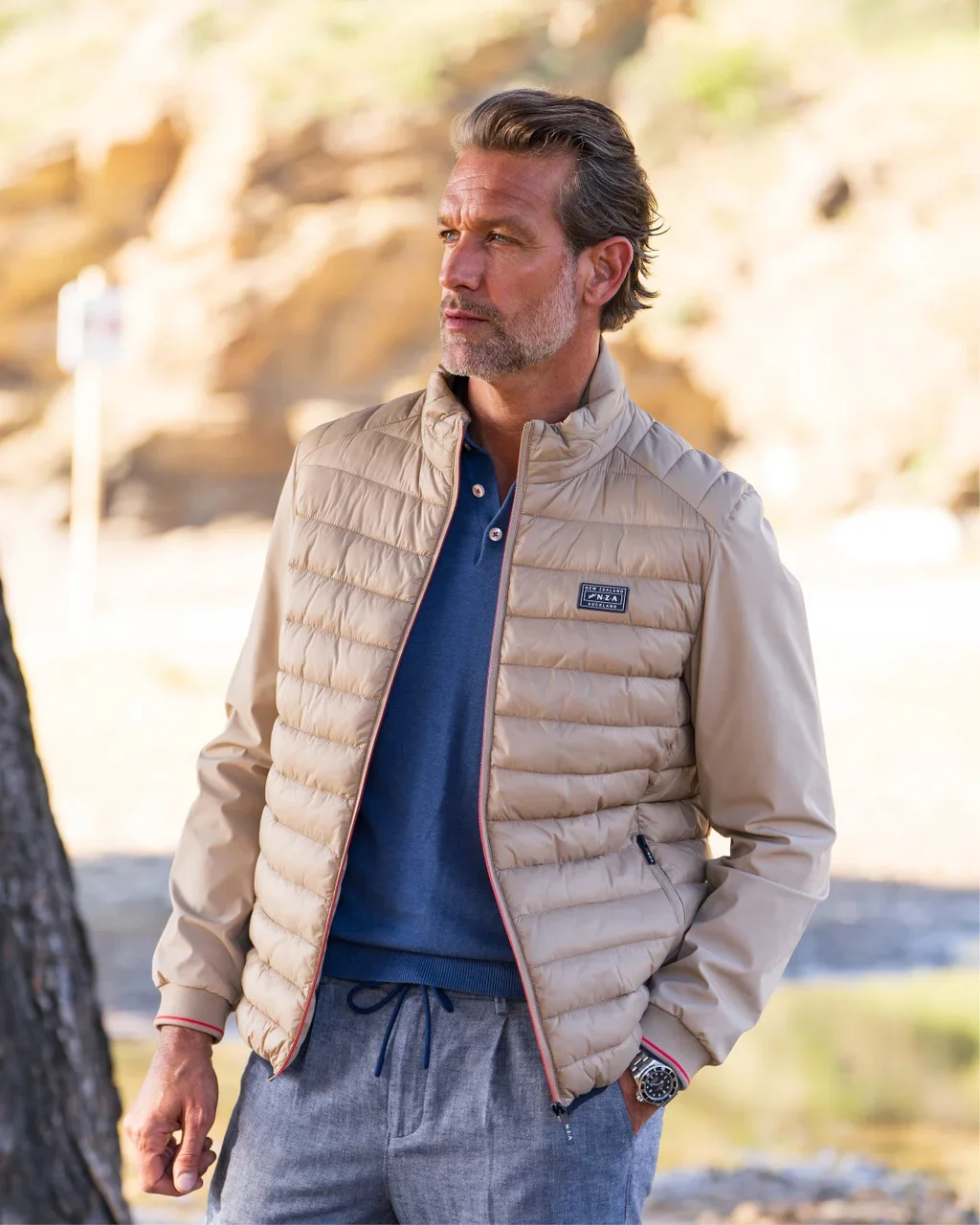 Man in beige puffer jacket over blue shirt and gray pants stands outdoors by a tree, looking to the side. Rocky background.