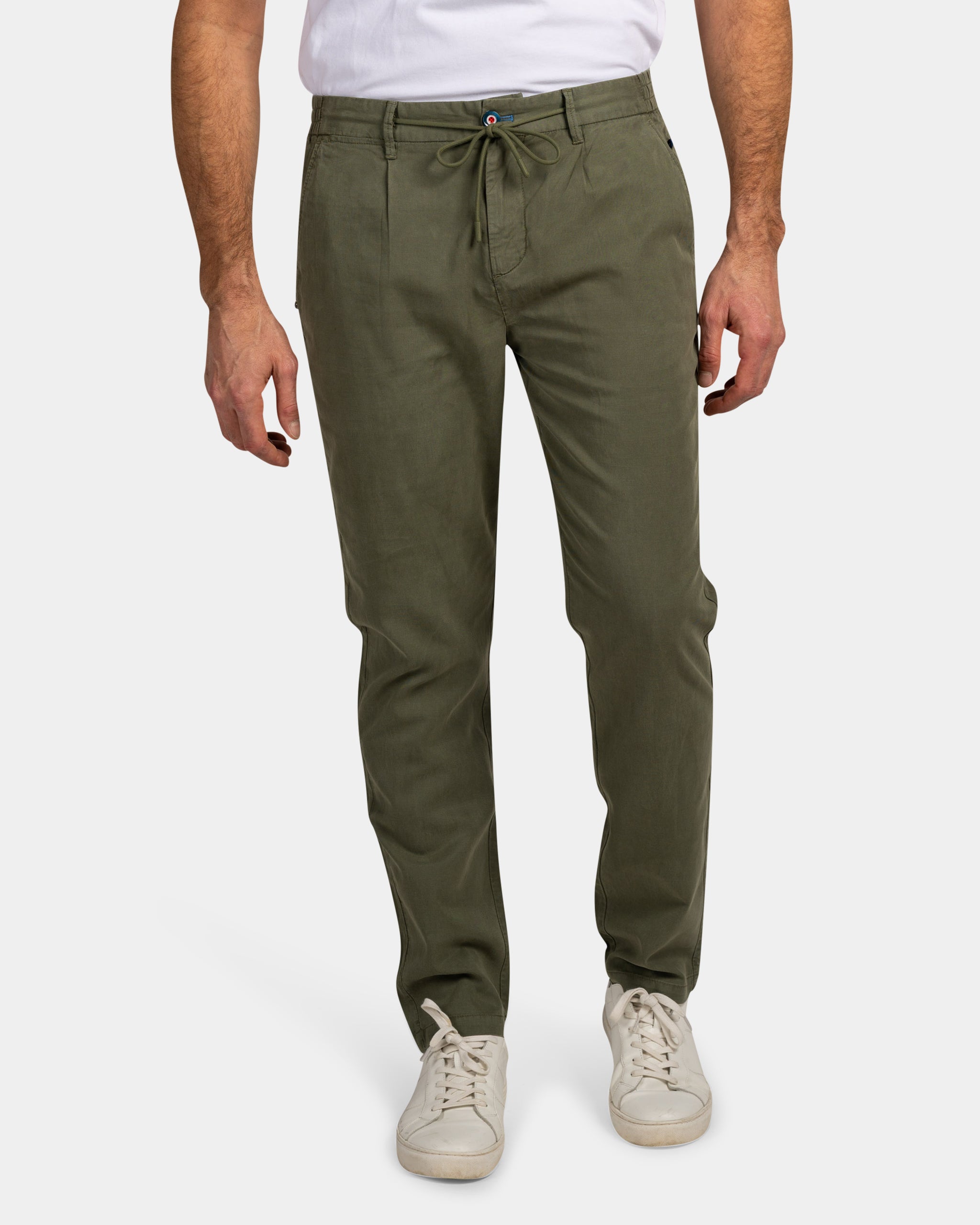 Man wearing olive green drawstring pants and white sneakers, hands resting by sides, against a plain white background.