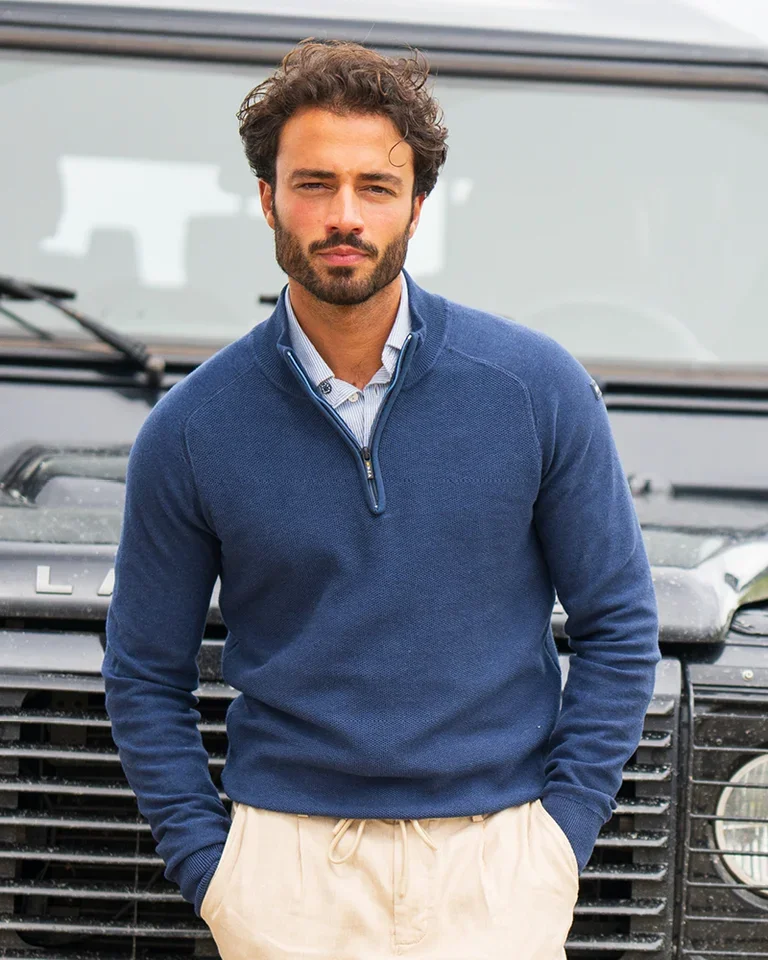 A man with a beard and short hair wears a blue quarter-zip sweater and beige pants, standing in front of a vehicle outdoors.