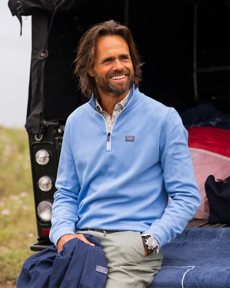 Smiling man with long hair sits on a vehicle tailgate, wearing a light blue pullover and holding a navy jacket.