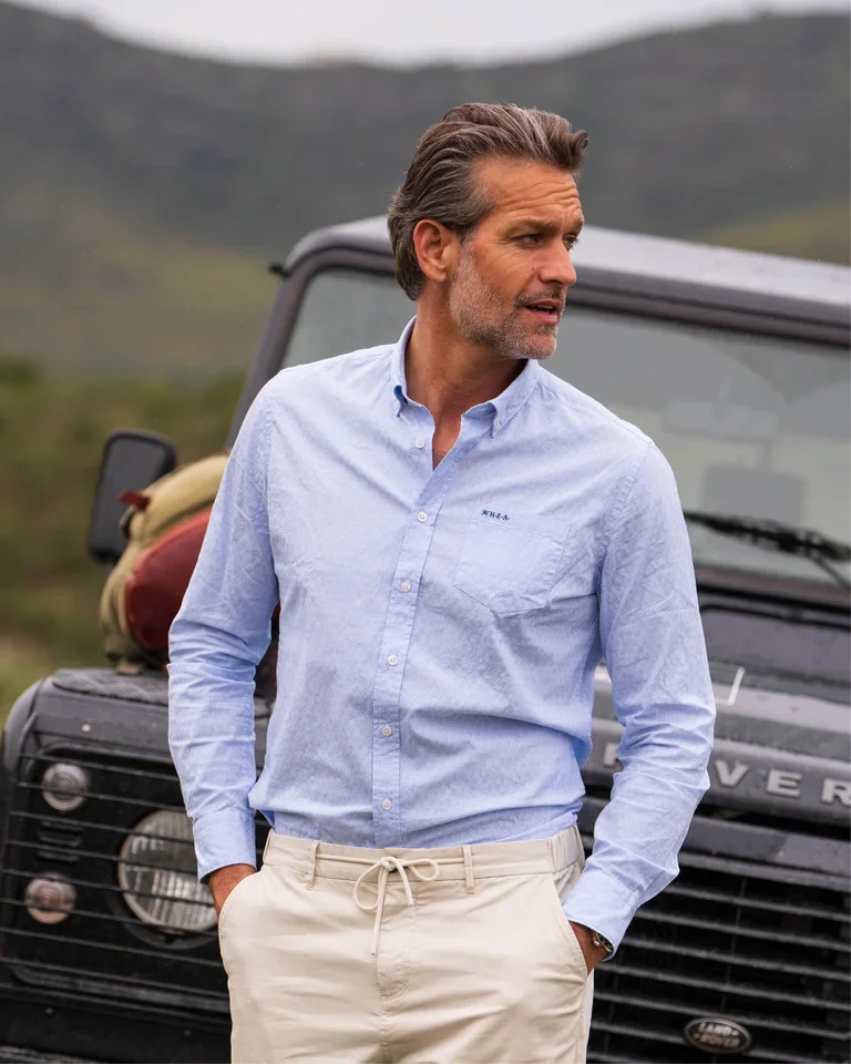 Man in light blue button-up shirt and beige pants stands in front of a black SUV in a mountainous area, looking to the side.