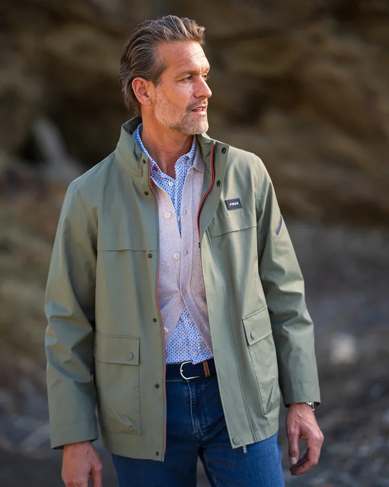Man in a green jacket and blue jeans standing outdoors, looking to the side. Background shows rocky terrain.