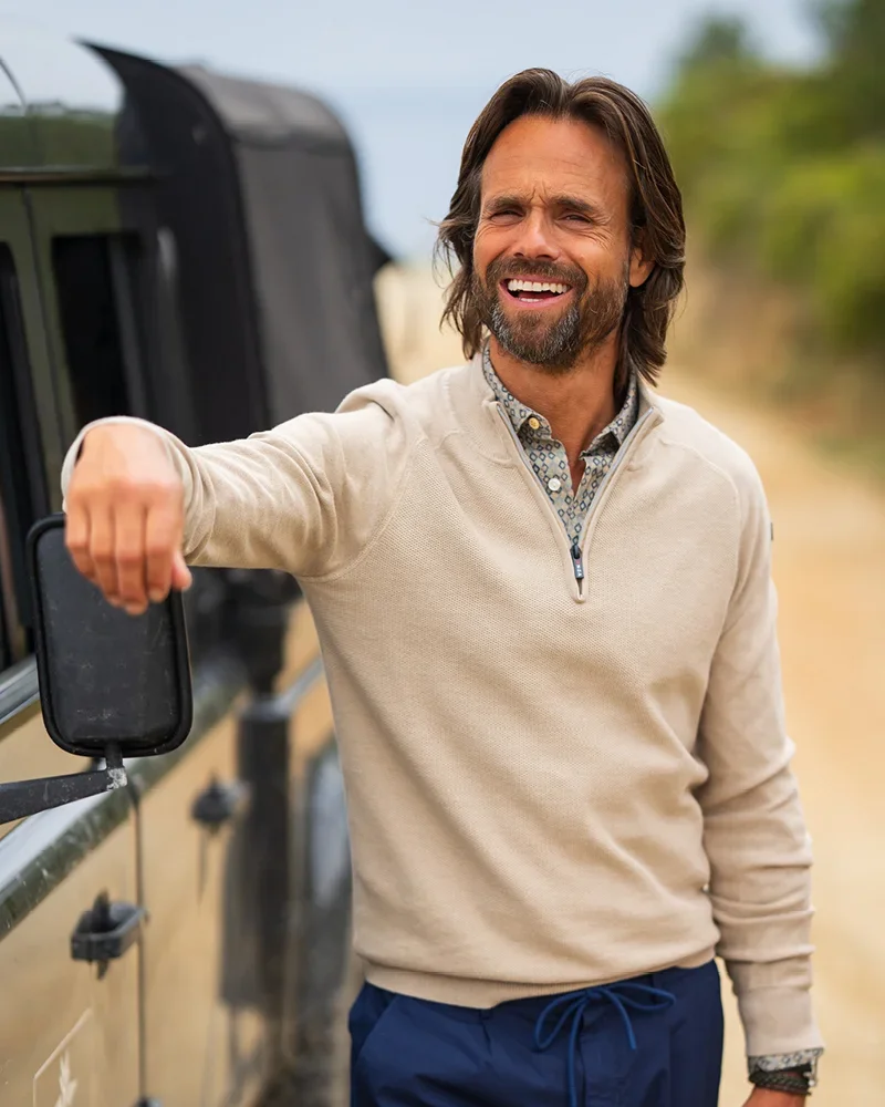 Man smiling, leaning on a vehicle, wearing a beige sweater and blue pants. Outdoor scene with blurred greenery in the background.