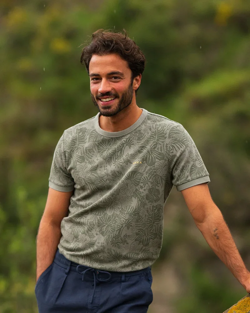 A man with a beard, wearing a green patterned T-shirt and blue pants, standing outdoors with a blurred green background, smiling.