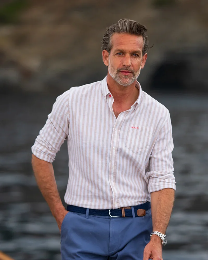 Man with salt-and-pepper hair wearing a striped shirt and blue pants stands outdoors near water, with rocky landscape in the background.